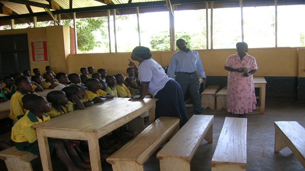 Ugandalainen alakoululuokka, jossa noin kaksikymmentä koulupukuista oppilasta istuu vierekkäin koulun penkeissä, ja opettaja seisoo heidän edessään luennoimassa.