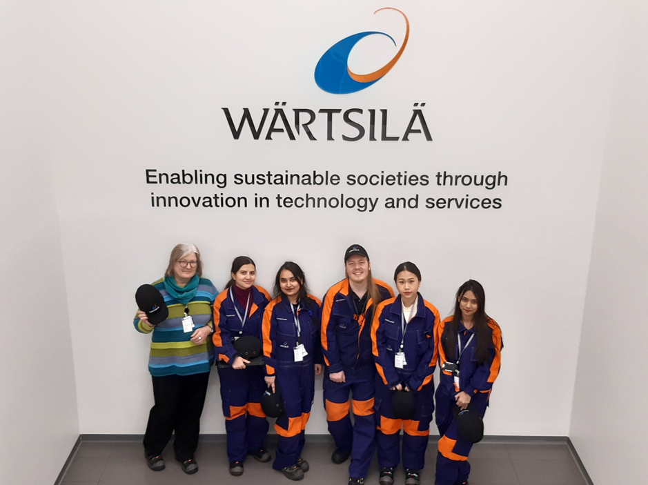 Kuvassa kaksi on ryhmä haalareihin pukeutuneita opiskelijoita opettajansa ja Wärtsilän kouluttajan kanssa portaikossa, jossa heidän takanaan seinällä on iso Wärtsilän logo ja tekstiä. Kuvassa on 5 naishenkilöä ja yksi mies. 