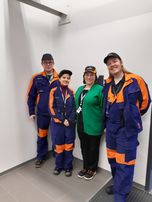 In this image number 6 we can see four persons who are wearing blue overalls and Wärtsiläs caps. There are two females and two males and they are all smiling. 