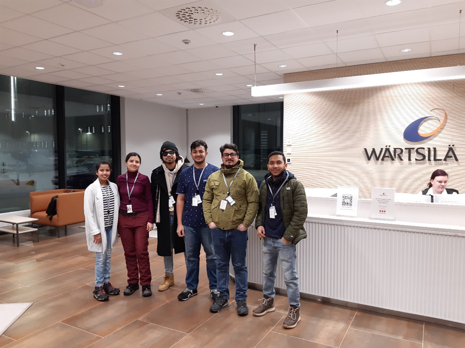 In this image number eight we can see a group of people standiing in the lobby of Wärtsilä´s factory STH building. There are two females and four males standing in front of a reception desk and one female worker is sitting behind the reception desk. Everybody are smiling. Wärtsilä logo can be seen on the wall behind the group. 