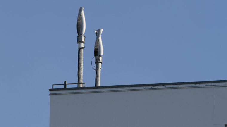 Kaksi pientuulivoimalaa on sijoitettu rakennuksen katolle. Tuulivoimalat ovat Savonius-tyyppisiä eli pystymallisia tuulivoimaloita. Tuulivoimalat ovat muutaman metrin pituisen maston päässä. Rakennus on valkoinen. Taustalla sininen taivas.