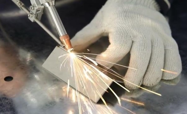 Manuaalista laserihitsausta G-Weike laitteella, jossa hitsauspistoolista suunnattu lasersäde kuumentaa levyn sulattaen sen. Kuvassa valkoinen hanskakäsi tukee hitsattavia ohuita metallilevyjä, jotka ovat hieman leveämpiä kuin sormi. Hitsauskipinät heijastuvat metallisesta työpöydästä.