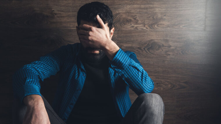 Mies istuu lattialla ja peittää kasvonsa kädellään.