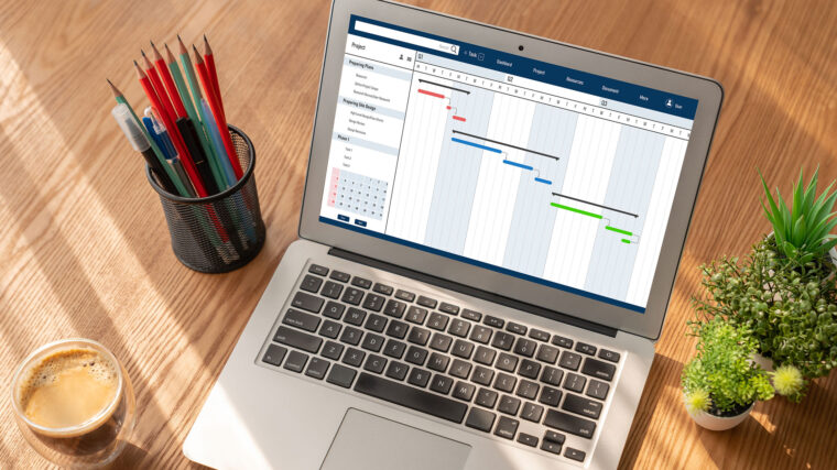 Computer showing a project planning software, Gantt charts. Pencils, coffee and plants on the table beside the laptop.