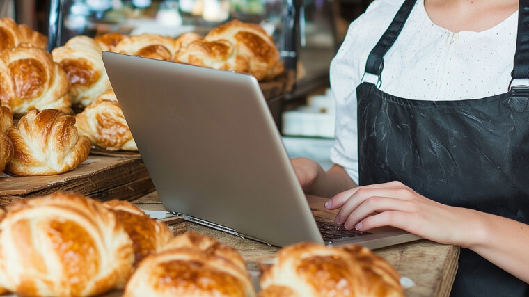 Nainen essu päällä, käyttää tietokonetta, ympärillä tuoreita leivonnaisia.