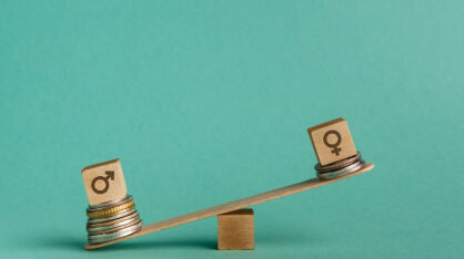 A seesaw with two wooden blocks on either end. The left block, marked with a male gender symbol, is supported by a tall stack of coins and sits lower. The right block, marked with a female gender symbol, is supported by a shorter stack of coins and sits higher, illustrating a gender pay gap.
