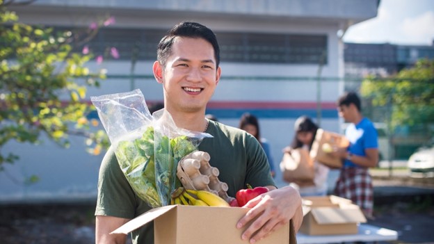Mies kulkee ruokatarvikkeita täynnä olevan pahvilaatikon kanssa iloisena.