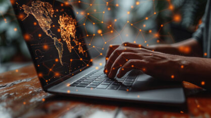 Lähikuva käsistä, jotka kirjoittavat kannettavan tietokoneen näppäimistöllä. Näytöllä näkyy digitaalinen maailmankartta, jossa on oransseja pisteitä ja viivoja, jotka muodostavat verkoston, symboloiden globaalia datayhteyttä ja teknologiaa.