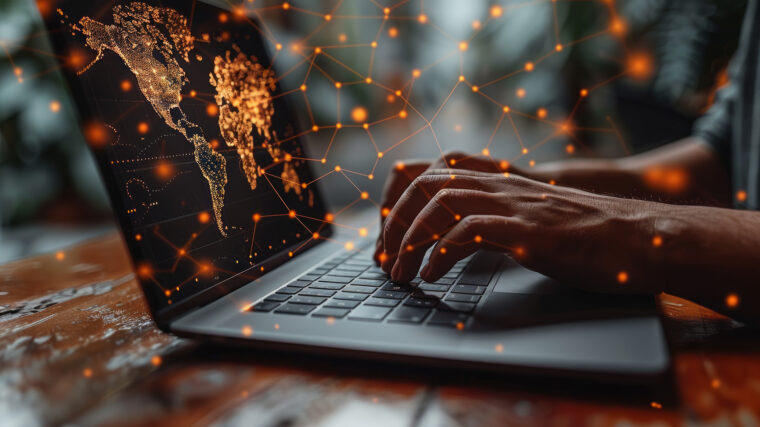 Lähikuva käsistä, jotka kirjoittavat kannettavan tietokoneen näppäimistöllä. Näytöllä näkyy digitaalinen maailmankartta, jossa on oransseja pisteitä ja viivoja, jotka muodostavat verkoston, symboloiden globaalia datayhteyttä ja teknologiaa.