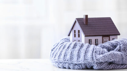 A small model house wrapped in a thick, light purple knitted scarf, symbolizing home warmth and insulation, placed on a white surface with a soft blurred background.