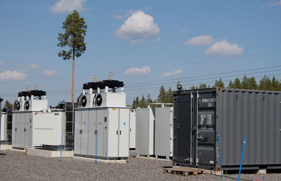 Kuvassa sähkövarasto Pyhännän Ainolassa. Etualalla sähkövarastoja sekä työmaavarasto. Taustalla yksittäinen mänty, metsänreuna, voimajohtolinja ja sinistä taivasta kesäisine pilvenhattaroineen.