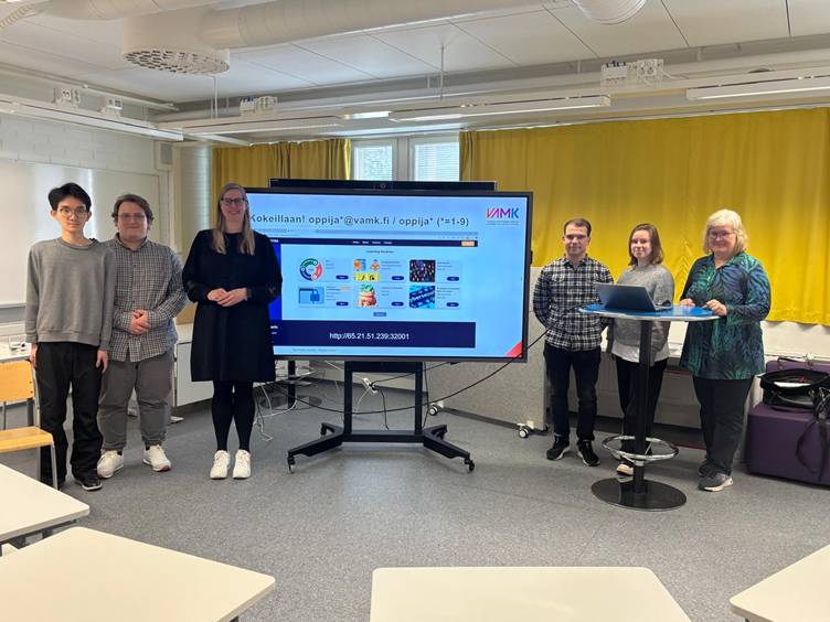 Henkilöitä seisomassa ison näytön molemmin puolin, näytöllä teksti "Kokeillaan!"