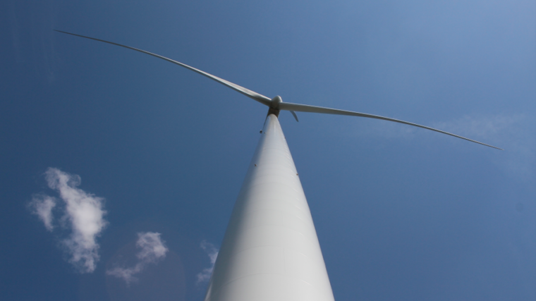 Kuvassa tuulivoimaturbiini alhaalta päin kuvattuna. Taustalla sininen taivas ja ohutta yläpilveä.