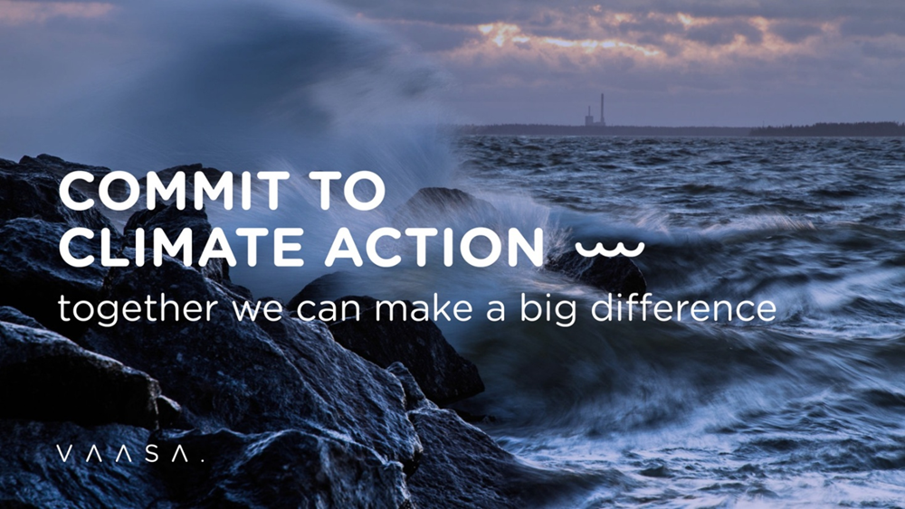 Rocky shoreline with waves crashing against the rocks under a cloudy sky at sunset. In the distance, an industrial structure is visible on the horizon. Overlaid text reads: “COMMIT TO CLIMATE ACTION — together we can make a big difference.” Bottom left corner shows the word “VAASA."
