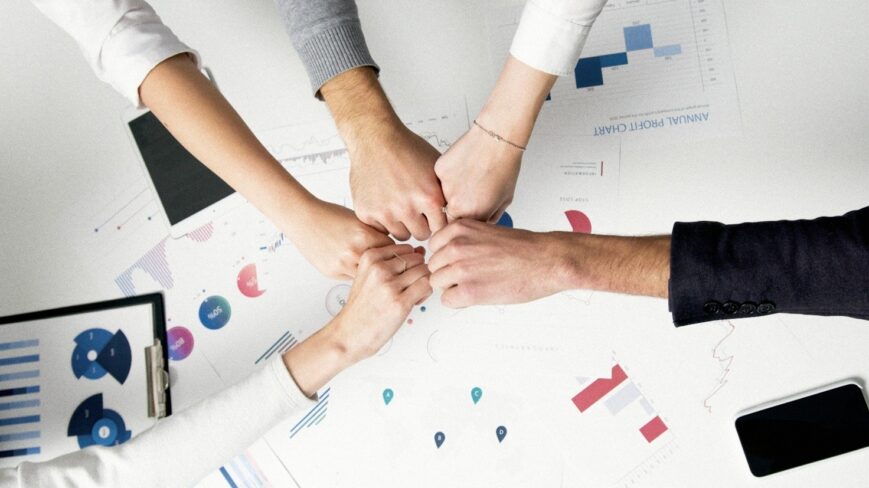 Neljä nyrkkiä yhdessä pöydän yläpuolella, pöydällä liiketoimintakaavioita ja älypuhelin.