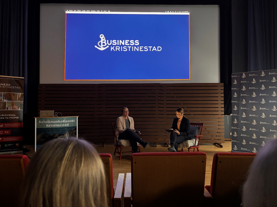 Kaksi ihmistä keskustelee keskenään istuen punaisilla tuolilla, etualalla katsomossa istuvia.