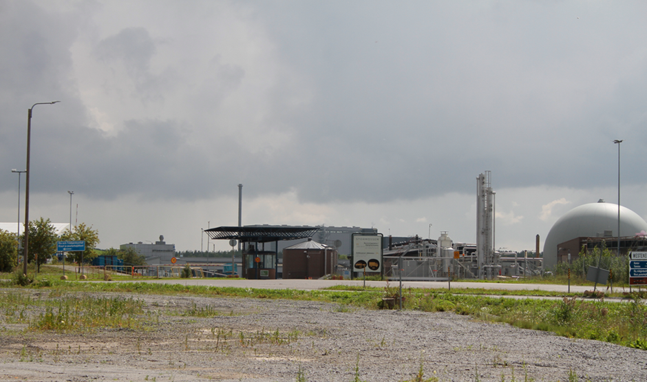 Kuvassa teollisuusalue, jolla sijaitsee biokaasulaitos. Etualalla näkymä matalaa heinä- ja rikkakasveja kasvavasta hiekkakentästä. Kuvan keskellä teollisuusalueelle johtava tie ja teollisuusalueen portti vartiorakennuksineen. Kuvassa näkyy myös biokaasulaitoksen kaasusäiliö ja soihtupolttopiippu epäpuhtaalle biokaasulle. Taivaalla tummia pilviä enteillen kesäistä ukkosta. 