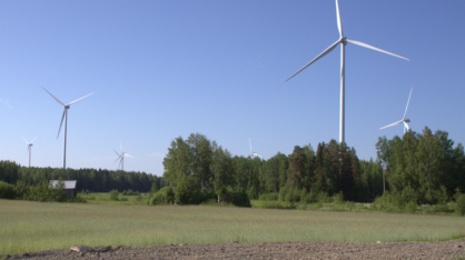 Kuvassa seitsemän tuulivoimaturbiinia maalaismaisemassa kesäkuun aamun valossa. Kuvan etualalla on kynnöspeltoa, jonka takana on kaistale heinäpeltoa. Tuulivoimapuiston edessä sijaitsee pellonreunan sekametsävyöhyke. Tuulivoimalat piirtyvät tarkkarajaisina vasten kesäaamun sinistä taivasta.