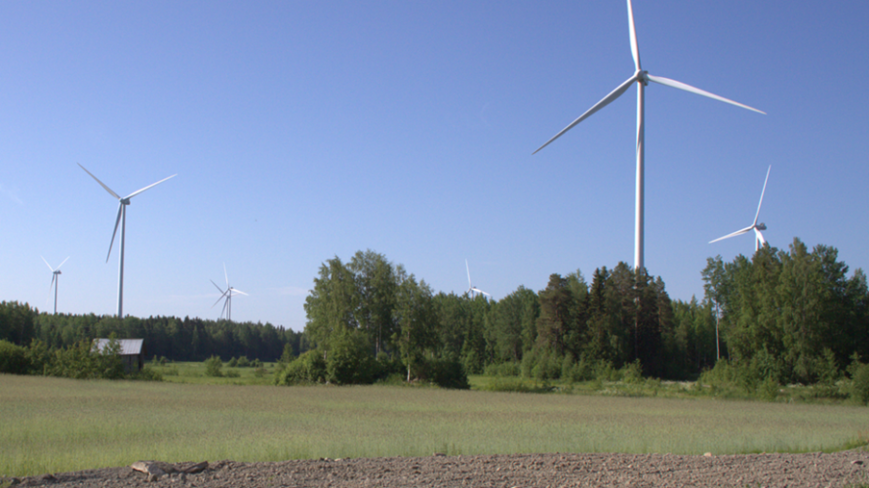 Kuvassa seitsemän tuulivoimaturbiinia maalaismaisemassa kesäkuun aamun valossa. Kuvan etualalla on kynnöspeltoa, jonka takana on kaistale heinäpeltoa. Tuulivoimapuiston edessä sijaitsee pellonreunan sekametsävyöhyke. Tuulivoimalat piirtyvät tarkkarajaisina vasten kesäaamun sinistä taivasta.