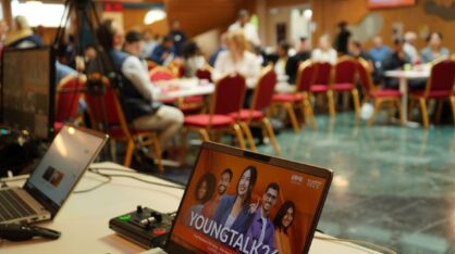 YoungTalk26-tapahtuma käynnissä: etualalla kannettava tietokone tapahtumaesityksellä, taustalla osallistujia istumassa pöytien ääressä.