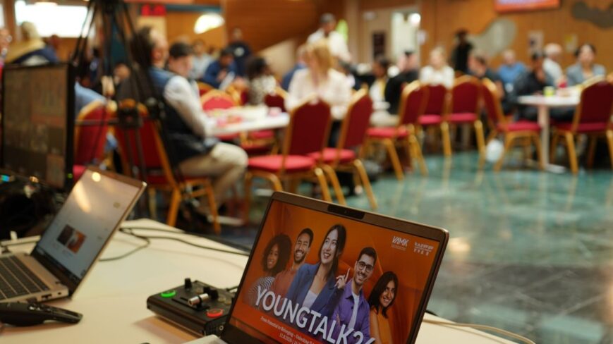 YoungTalk26-tapahtuma käynnissä: etualalla kannettava tietokone tapahtumaesityksellä, taustalla osallistujia istumassa pöytien ääressä.